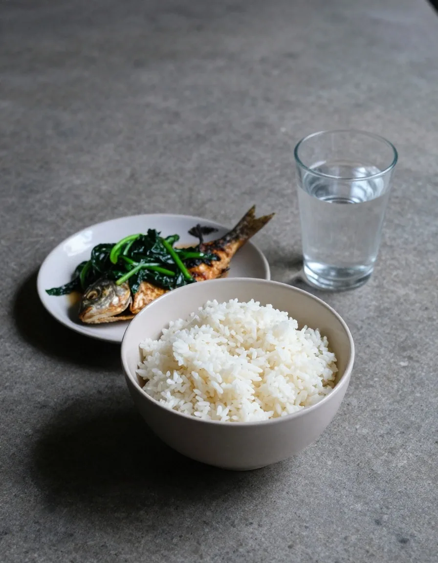 A simple rice, fish and greens meal with a glass of water on a stone counter